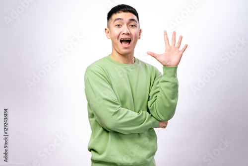 An Asian man in a green sweatshirt is smiling while holding up five fingers on his right hand. His left arm is crossed, and he stands against a white background, conveying a friendly photo