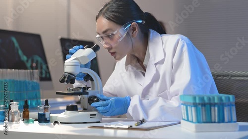 A woman in a lab coat is looking through a microscope. She is wearing blue gloves and has a serious expression on her face. The lab is filled with various scientific equipment and materials