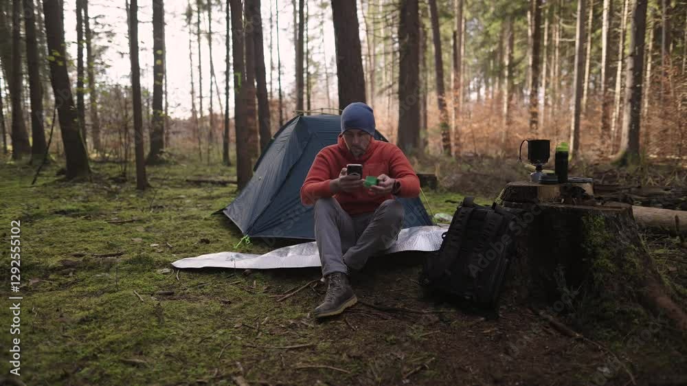 Male hiker drinking tea and surfing Internet on smartphone in tent camp during hiking in fall in woods. Backpacker makes route in map in phone and drinks hot drink from cup in background of tent. 