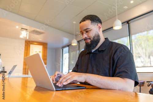 An African American man focuses on his laptop at a wooden table in a bright coffee shop, where natural light streams through large windows, creating a cozy atmosphere for creativity