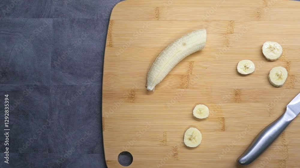 A partially peeled banana lies on a wooden cutting board with several freshly sliced pieces beside it. A knife is placed nearby, indicating food preparation.