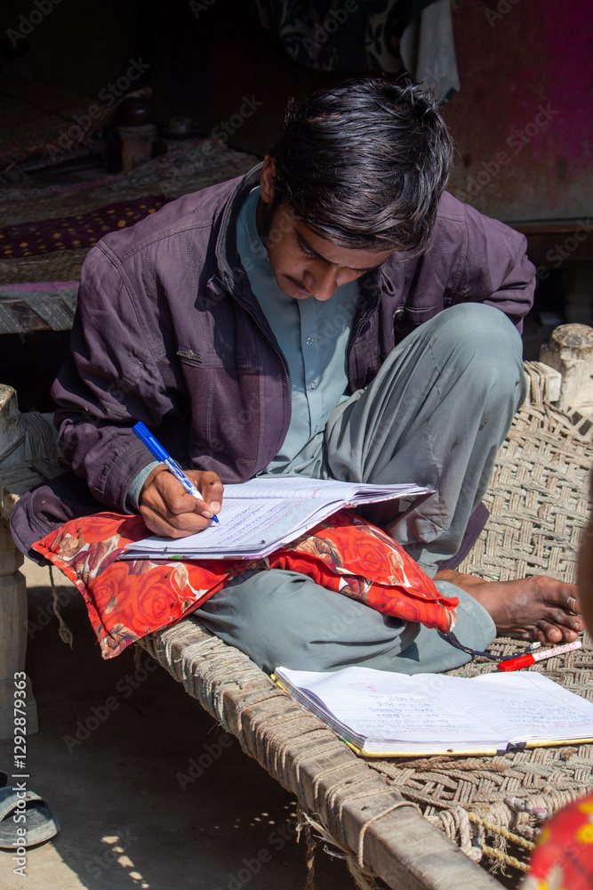 Fototapeta premium Pakistani Village Boy Studying on a Charpai | Rural Education Concept | Focused Young Student Writing in Notebook Outdoors