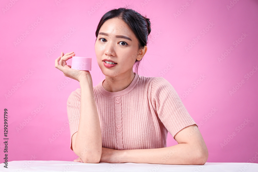 A young Asian woman with dark hair in a bun, wearing a ribbed pink short-sleeve top, holds a pink skincare cream jar while touching her face, promoting skincare against soft pink background