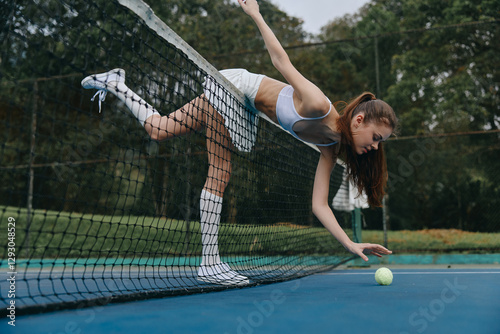 Young female tennis player bending over the net to retrieve a ball on a sunny court, showcasing determination and athleticism in stylish sport attire