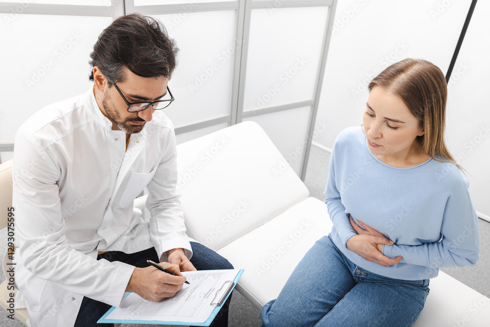 Woman with abdominal pain during a consultation with gastroenterologist who checks condition of her internal organs, such as stomach, liver, gallbladder, pancreas, and intestines Woman with abdominal pain during a consultation with gastroenterologist who checks condition of her internal organs, such as stomach, liver, gallbladder, pancreas, and intestines