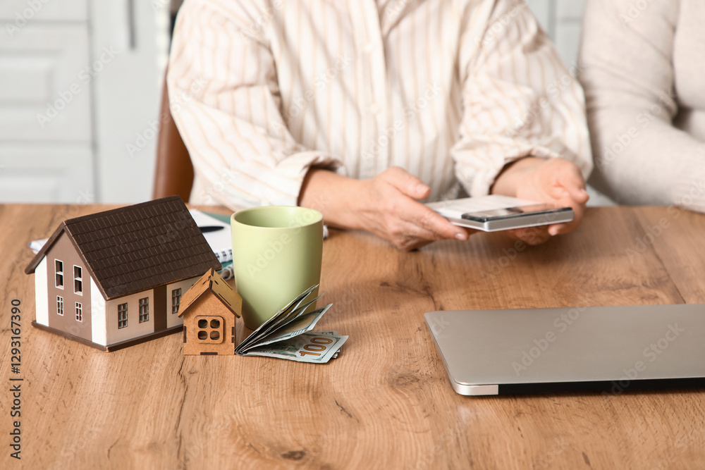 House figures with money on table against senior couple calculating utility bills in kitchen, closeup House figures with money on table against senior couple calculating utility bills in kitchen, closeup