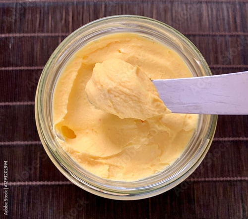 A glass jar filled with creamy yellow butter sits on a wooden surface, while a knife reveals its smooth and spreadable texture, inviting culinary use in various dishes