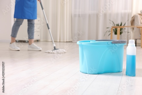 Woman cleaning floor with string mop indoors, focus on bucket and bottle of detergent