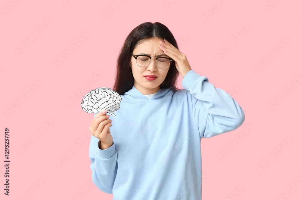 Young Asian woman with paper brain suffering from headache on pink background. Brain Injury Awareness Month Young Asian woman with paper brain suffering from headache on pink background. Brain Injury Awareness Month