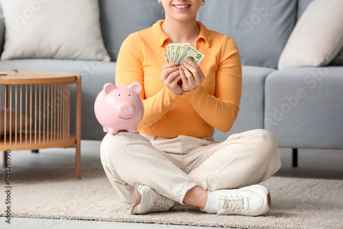 Young woman with piggy bank and dollar banknotes sitting on floor at home