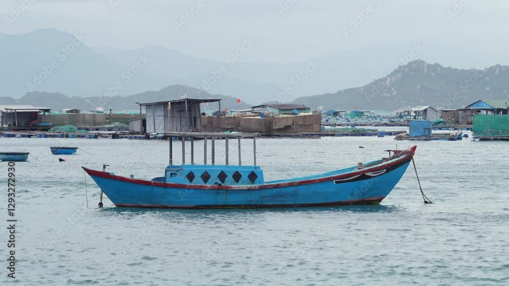 A traditional blue fishing boat floats on calm waters near a floating village, with mountains and fishing structures in the background. A traditional blue fishing boat floats on calm waters near a floating village, with mountains and fishing structures in the background.