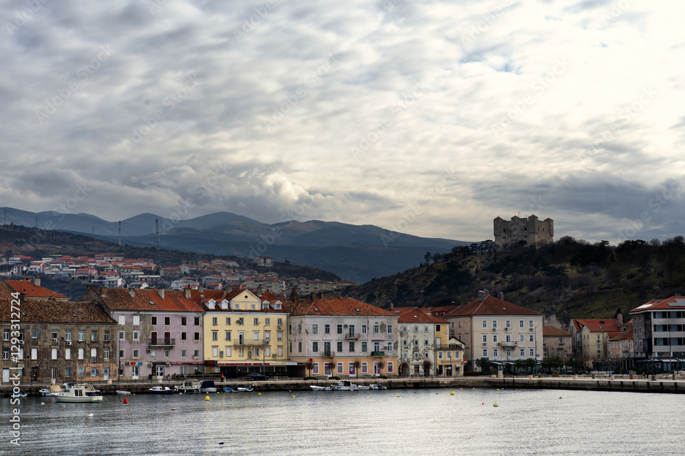 Obraz premium Senj, Croatia, showcases its historic charm with colorful buildings along the waterfront, a hilltop castle, and rolling hills under a dramatic, cloudy sky.