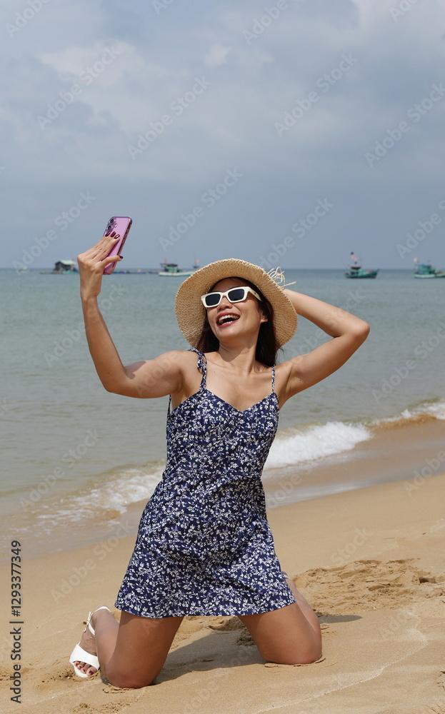 Asian woman wearing a sleeveless, floral-patterned short dress snaps selfies kneeling on a sandy beach Asian woman wearing a sleeveless, floral-patterned short dress snaps selfies kneeling on a sandy beach