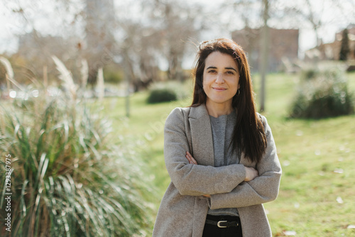A confident woman in a stylish coat stands outdoors with arms crossed, exuding self-assurance and poise. The blurred natural background enhances depth and focus. Horizontal, copy-space