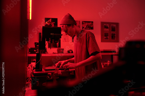 Moody side view portrait of teenage boy working with equipment in darkroom while developing photo prints in red light young photographer in studio copy space