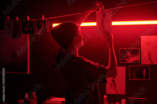 Red light image of photographer working with traditional media and hanging developed photo print on drying line under red neon light in darkroom