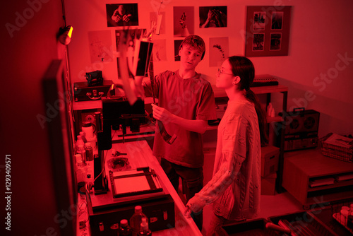 Teenage boy discussing developed photo prints and doing selections while working together in darkroom lit by red light young photographers in studio copy space photo