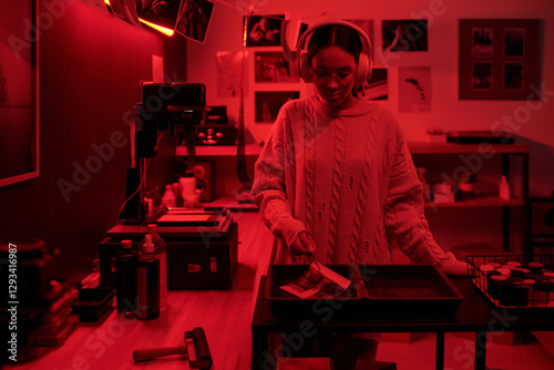 Waist up portrait of teenage girl wearing headphones developing analog photos in trays working in darkroom lit by red light photographer in studio copy space