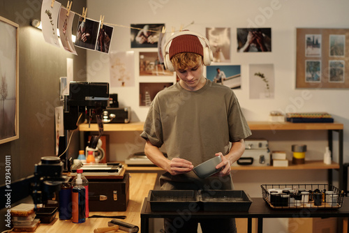 Front view portrait of young photographer mixing chemicals while developing traditional photography in darkroom and wearing headphones, copy space