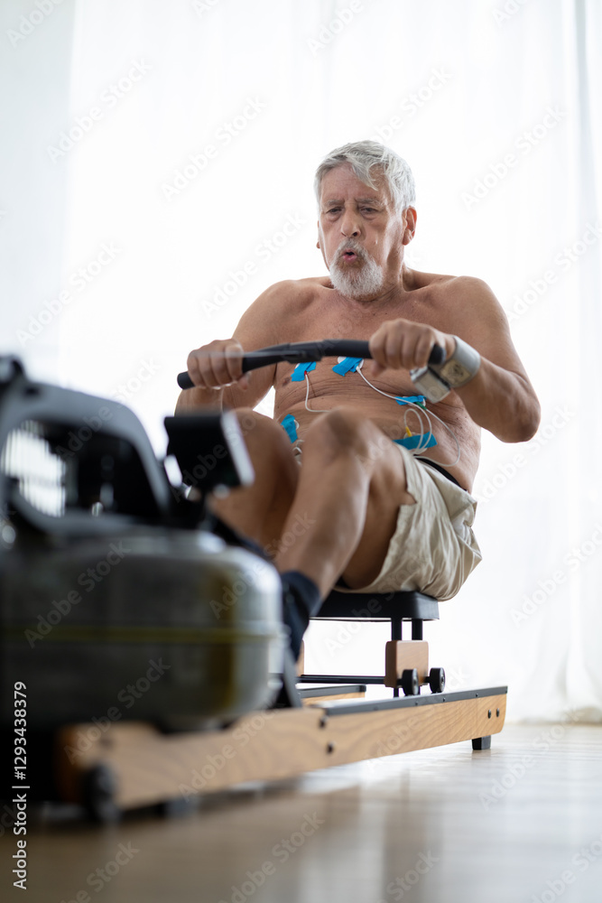 Staying fit and healthy. Senior shirtless man with an EKG mobile device exercising on rowing a machine at home, measuring his heart rate, pressure and pulse.