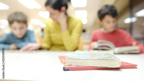 Books in foreground, blurred: kids and woman reading/studying. Warm, soft lighting. Library/classroom setting.