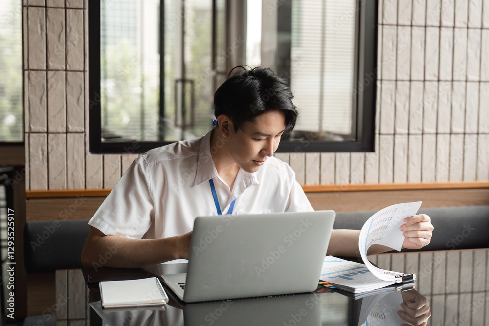 Young professional analyzing documents while working on laptop in modern office, focusing on data and project details. Young professional analyzing documents while working on laptop in modern office, focusing on data and project details.