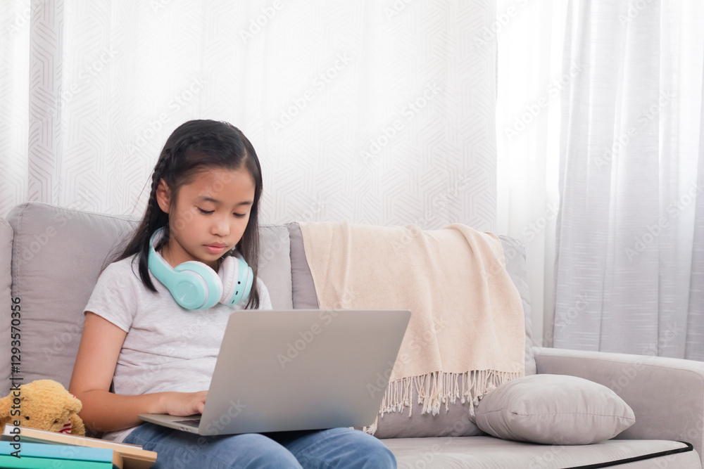 Asian young girl using laptop on sofa with headphones around neck. Focused on screen, possibly engaged in online learning or entertainment. Cozy living room setting with teddy bear and books. Asian young girl using laptop on sofa with headphones around neck. Focused on screen, possibly engaged in online learning or entertainment. Cozy living room setting with teddy bear and books.
