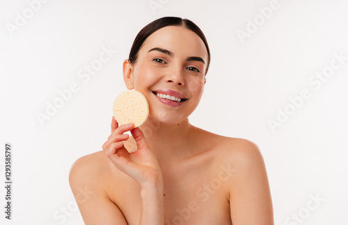 Person with bright smile holds a yellow facial sponge against clear skin in minimalist setting. Portrays concepts of skincare, happiness, and natural beauty. Enhances sense of well-being and radiance.