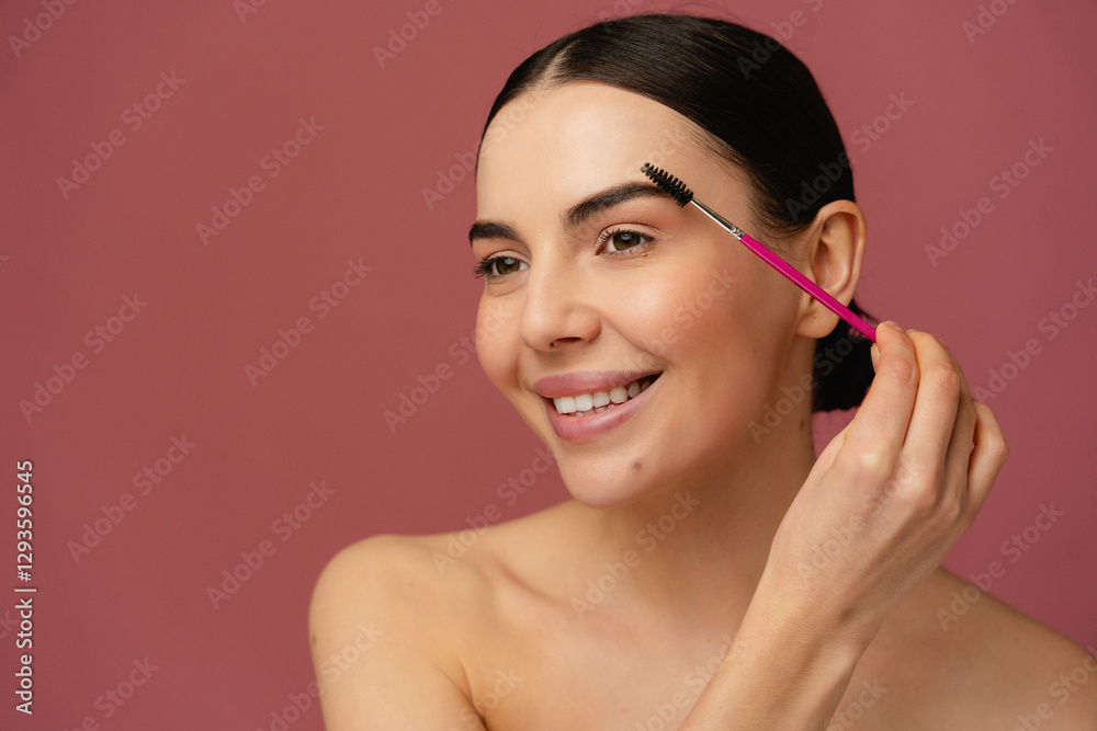Close-up of a cheerful individual enhancing eyebrows with a grooming brush, set against a pink backdrop, depicting personal care and beauty practices. Close-up of a cheerful individual enhancing eyebrows with a grooming brush, set against a pink backdrop, depicting personal care and beauty practices.