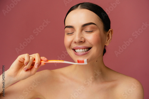 Close-up of a girl smiling and brushing teeth with an orange toothbrush, set against a soft pink backdrop. Highlights the concept of oral hygiene, self-care, and positivity.