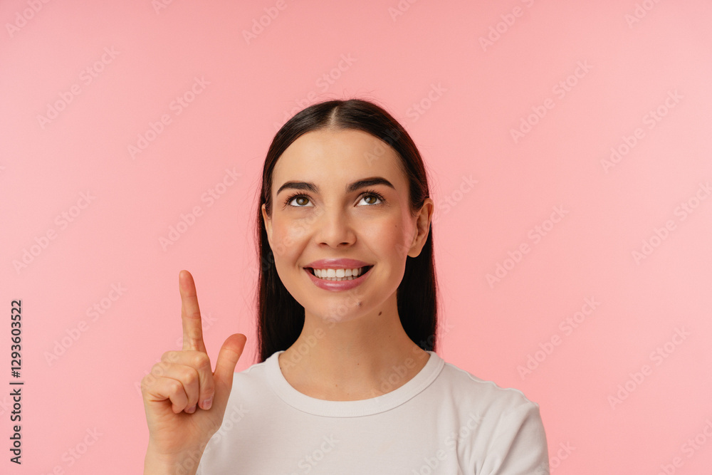 Cheerful person looking upward and pointing with a bright smile, standing against a pink background. Cheerful person looking upward and pointing with a bright smile, standing against a pink background.