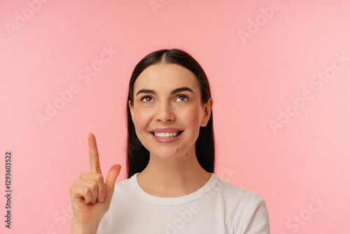 Cheerful person looking upward and pointing with a bright smile, standing against a pink background.