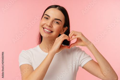 Happy beautiful female model girl wearing a white shirt shows a heart symbol with hands while smiling warmly against a colorful pink background.