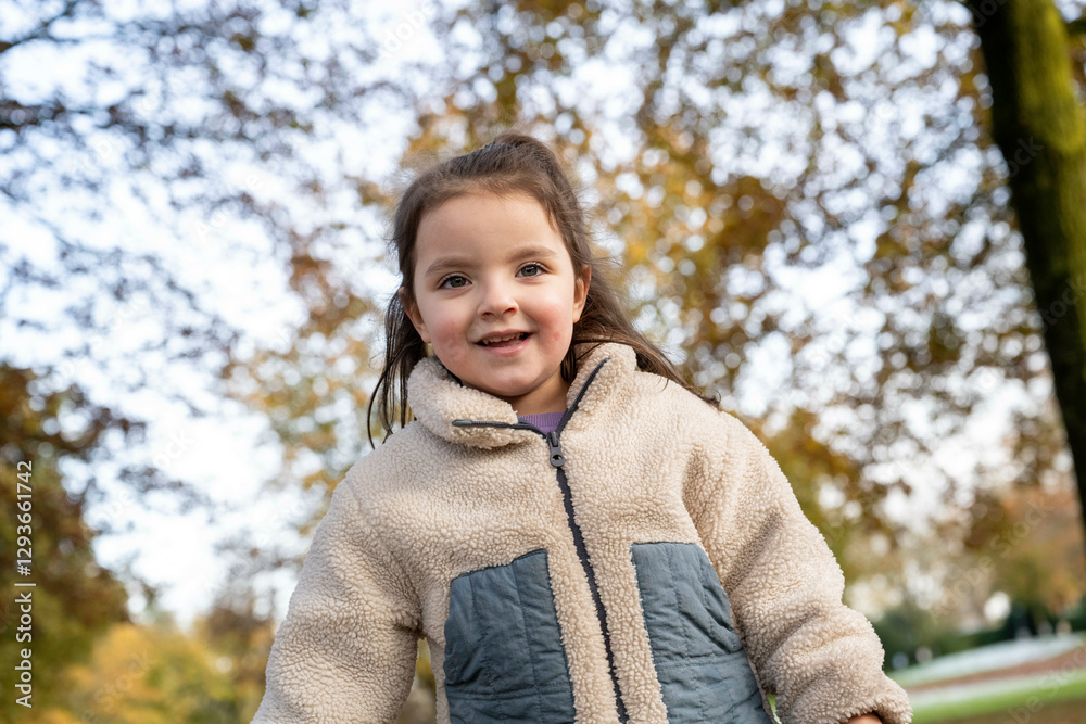 Smiling young girl in a cozy jacket standing outdoors on an autumn day with trees and fallen leaves in the background, Netherlands Smiling young girl in a cozy jacket standing outdoors on an autumn day with trees and fallen leaves in the background, Netherlands