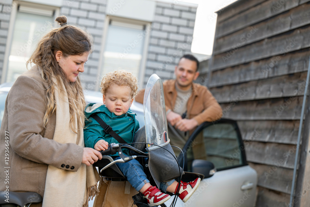 A smiling mother with a toddler on a bike and father leaning on a car outside their house, Netherlands A smiling mother with a toddler on a bike and father leaning on a car outside their house, Netherlands