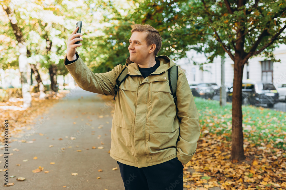 Obraz premium Smiling 30s man with backpack taking photo on park background. Selfie time. Holidays, rest, blogging banner. Charming male wearing stylish outfit have fine