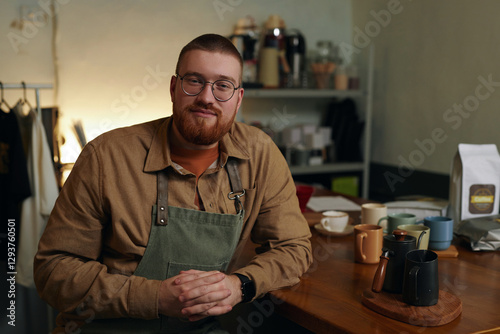 Medium close up portrait of Caucasian young adult entrepreneur smiling while sitting at table with handmade cups for coffee photo