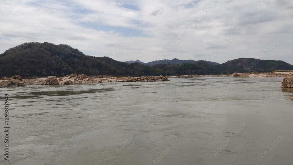 Shallow river water reflecting the cloudy sky, with fine sand patterns in the foreground. Rocky riverbanks and distant mountains create a serene natural landscape, perfect for travel, nature, and envi Shallow river water reflecting the cloudy sky, with fine sand patterns in the foreground. Rocky riverbanks and distant mountains create a serene natural landscape, perfect for travel, nature, and envi