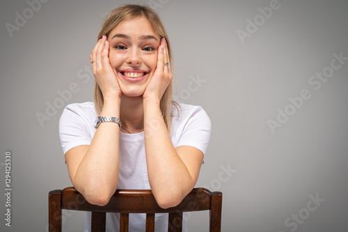 Happy, emotional young girl with blond hair smiling and leaning on wooden chair on neutral background. Cheerful woman in casual white t-shirt, confidence, joy, positivity, space for inscription