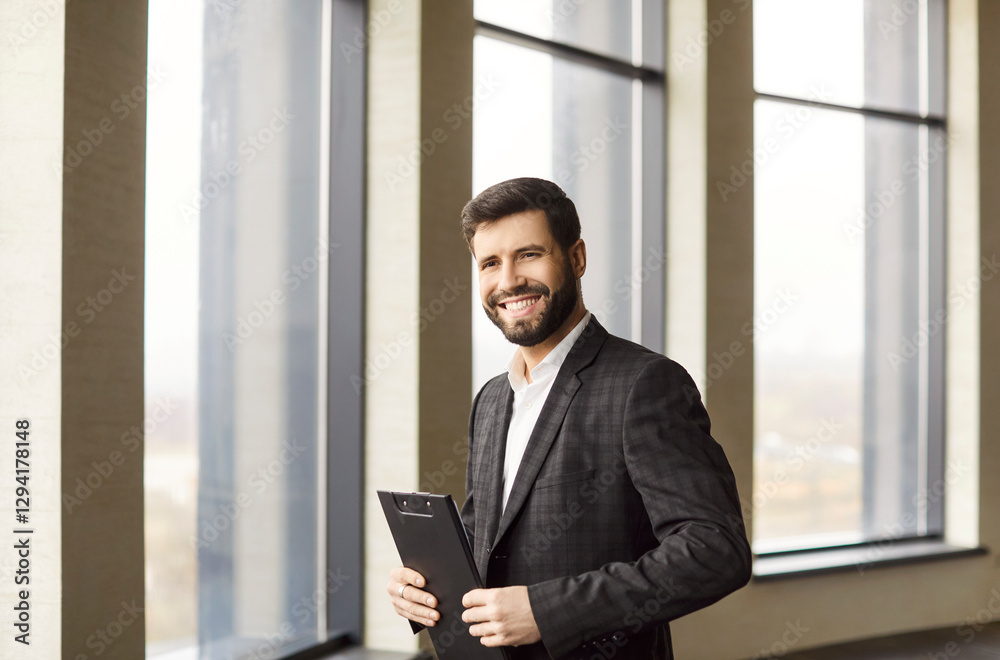 Young smiling attractive entrepreneur in formal suit standing in modern office. Real estate agent man holding documents and looking cheerful at camera. Business portrait and real estate concept. Young smiling attractive entrepreneur in formal suit standing in modern office. Real estate agent man holding documents and looking cheerful at camera. Business portrait and real estate concept.