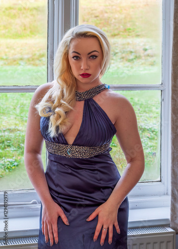 Beautiful woman in blue evening dress in front of window staring at observer