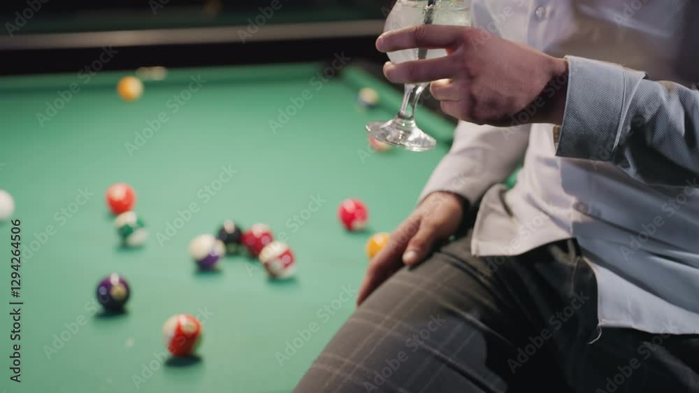 Person in white shirt and grey trousers holding lemon drink with black straw, sitting on green pool table with colorful billiard balls. Casual setting, stylish attire, dim ambiance, and relaxed mood