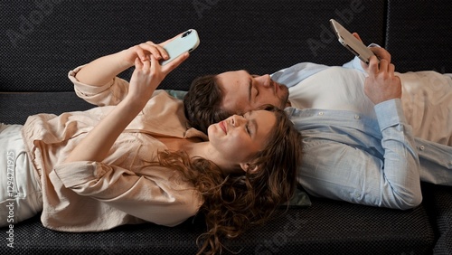 Lovely couple relaxing on the couch at home, man and woman laying on the sofa and scrolling social media online on smartphones.