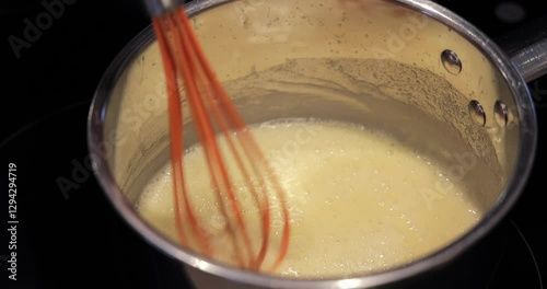Warm milk is poured into melted butter in a saucepan while a whisk stirs the mixture creating the base for bechamel sauce in a close-up cooking process shot