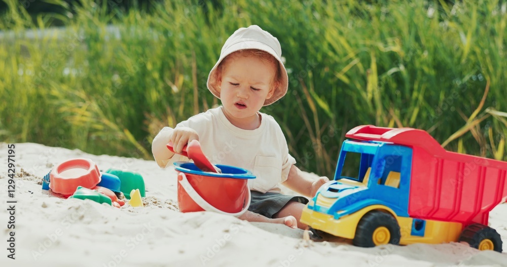 Boy on the beach near the water pours sand into a bucket with a shovel, surrounded by toys, deeply focused on the activity. Boy on the beach near the water pours sand into a bucket with a shovel, surrounded by toys, deeply focused on the activity.