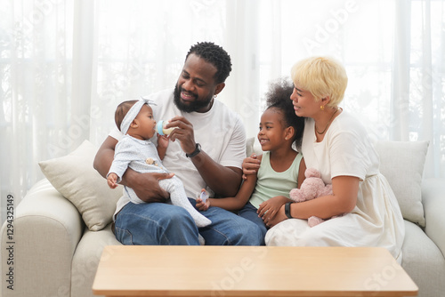 Loving interracial family sitting on couch at home,an african father take care a baby,feeding milk and asian mother playing with a girl