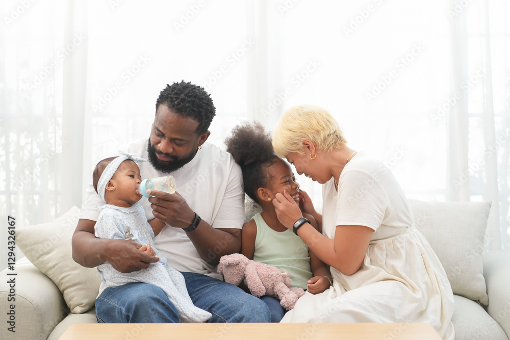 Loving interracial family sitting on couch at home,an african father take care a baby,feeding milk and asian mother playing with a girl Loving interracial family sitting on couch at home,an african father take care a baby,feeding milk and asian mother playing with a girl