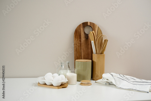 A curated collection of kitchen utensils and fresh ingredients: eggs, milk, sugar, cutboard, napkin is elegantly displayed on a neutral white table. photo