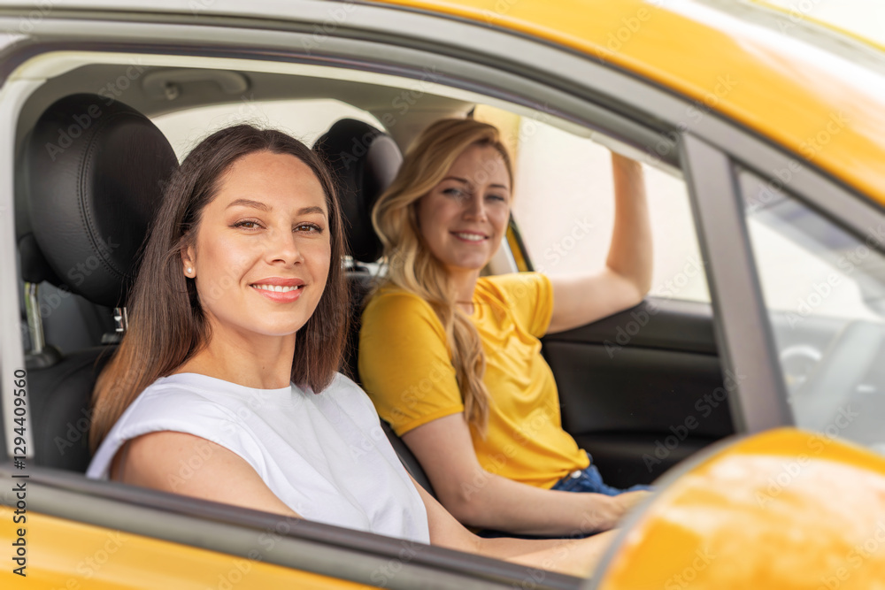 Two smiling women sit inside a yellow taxi, one in the drivers seat and the other in the passengers seat, enjoying their ride.