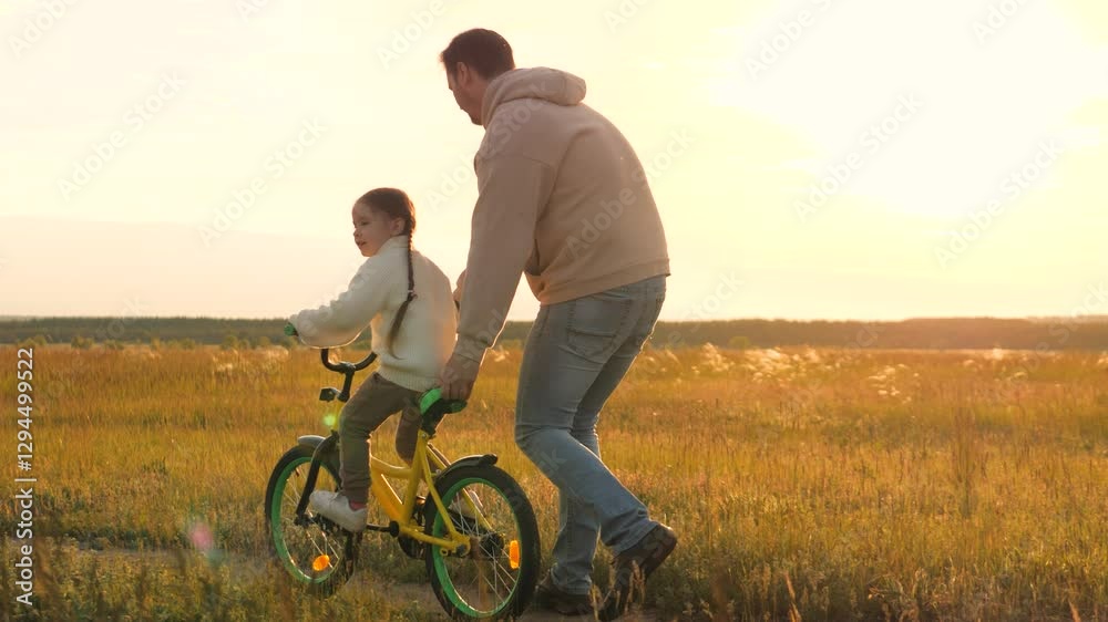 Parent teaches child to ride bicycle. Father and daughter in field at epic sunset. Child safety, dad regulates and controls beloved girl kid on bike, warning against injuries and bruises. Slow motion Parent teaches child to ride bicycle. Father and daughter in field at epic sunset. Child safety, dad regulates and controls beloved girl kid on bike, warning against injuries and bruises. Slow motion